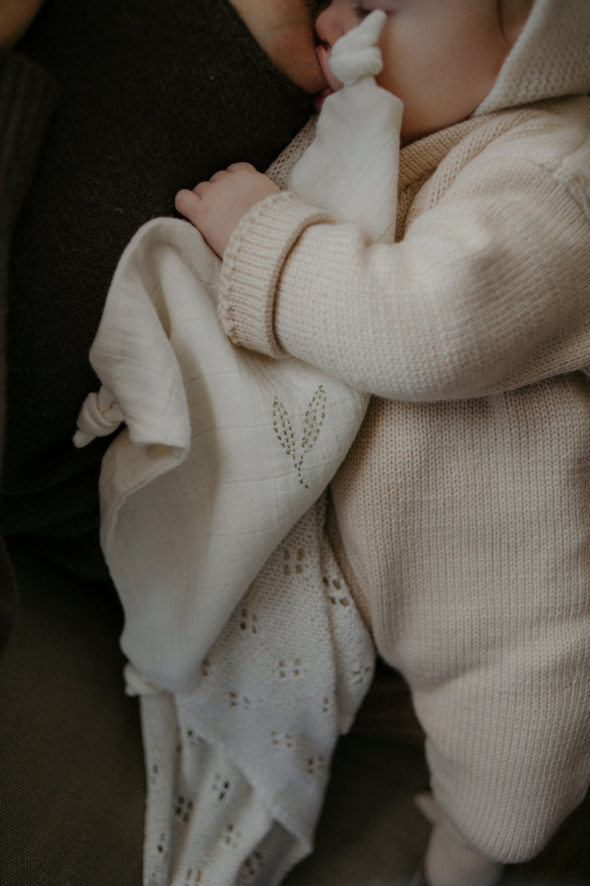 Baby in a beige knit outfit holding a cream organic cotton comforter with lace details.