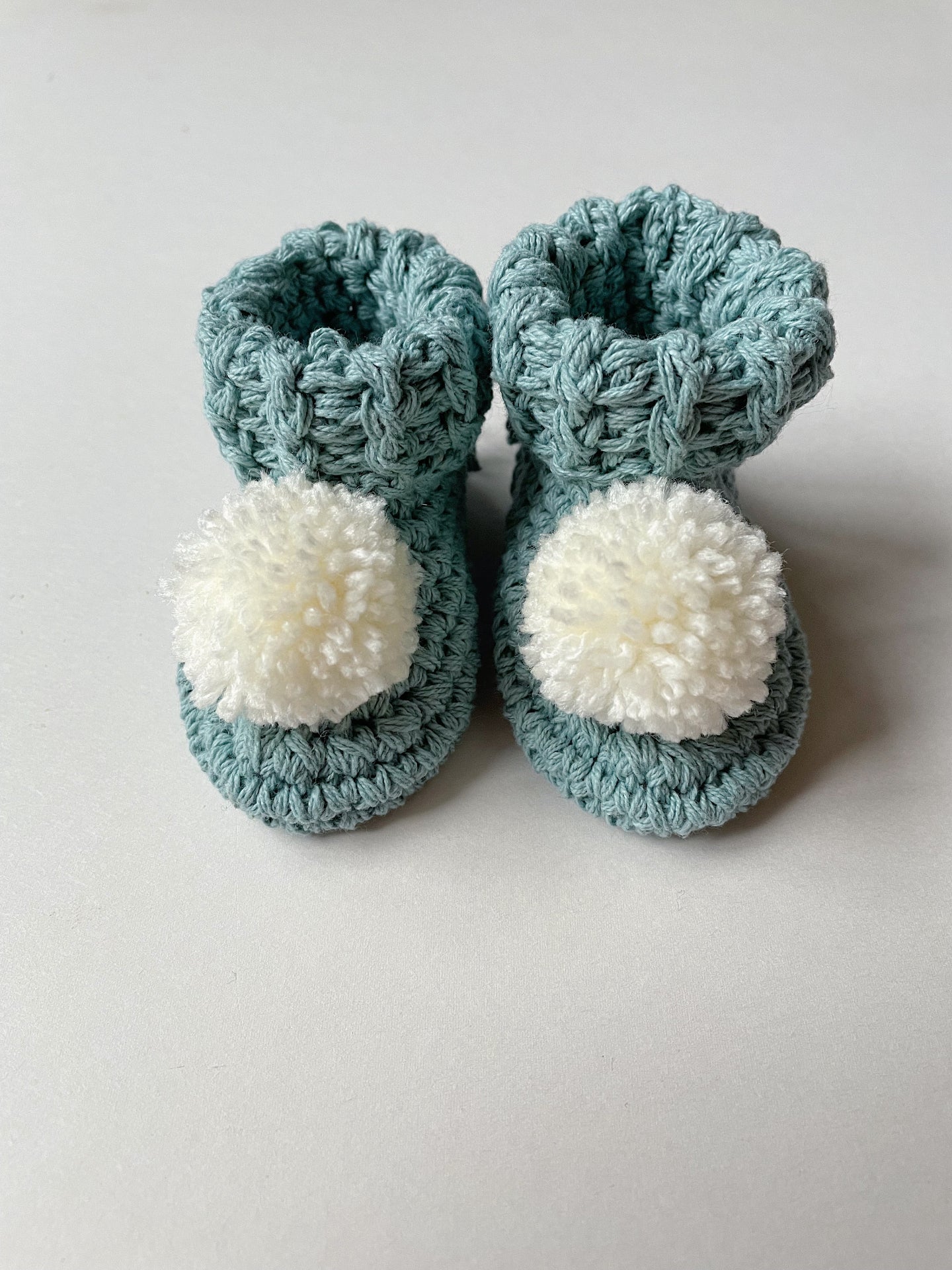 image shows a pair of duck egg coloured organic cotton baby booties with cream pom poms