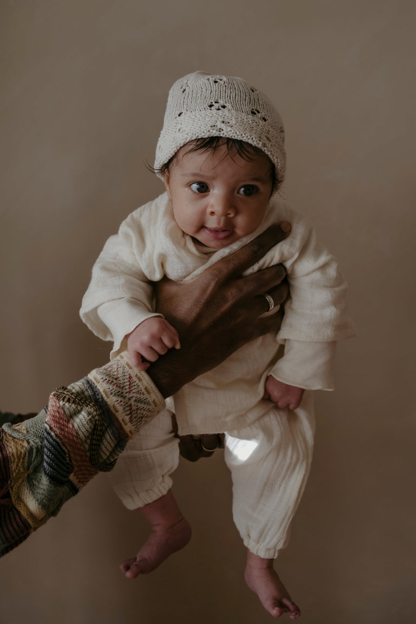 Baby in cream organic cotton pyjamas and knit hat being held against a plain background