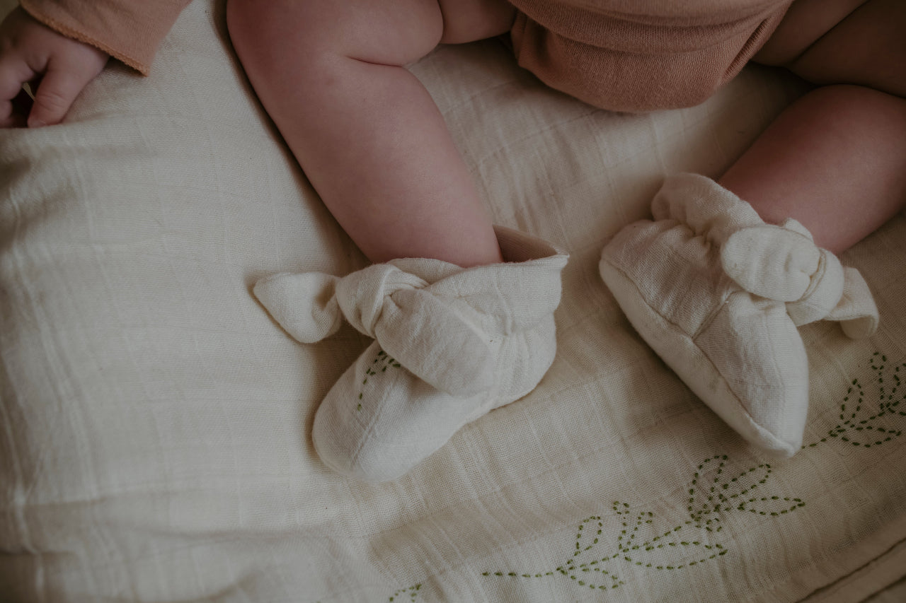 Baby's feet wearing cream organic cotton booties with bows on a textured surface