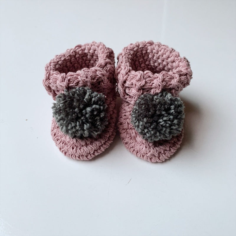 image shows a pair of pink baby booties with a dark grey pom pom