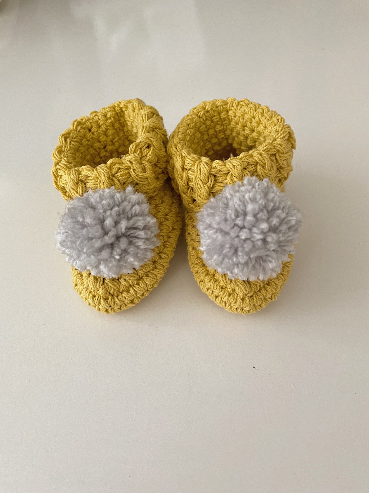 Image shows a pair of mustard yellow cotton baby booties with light grey pom pom